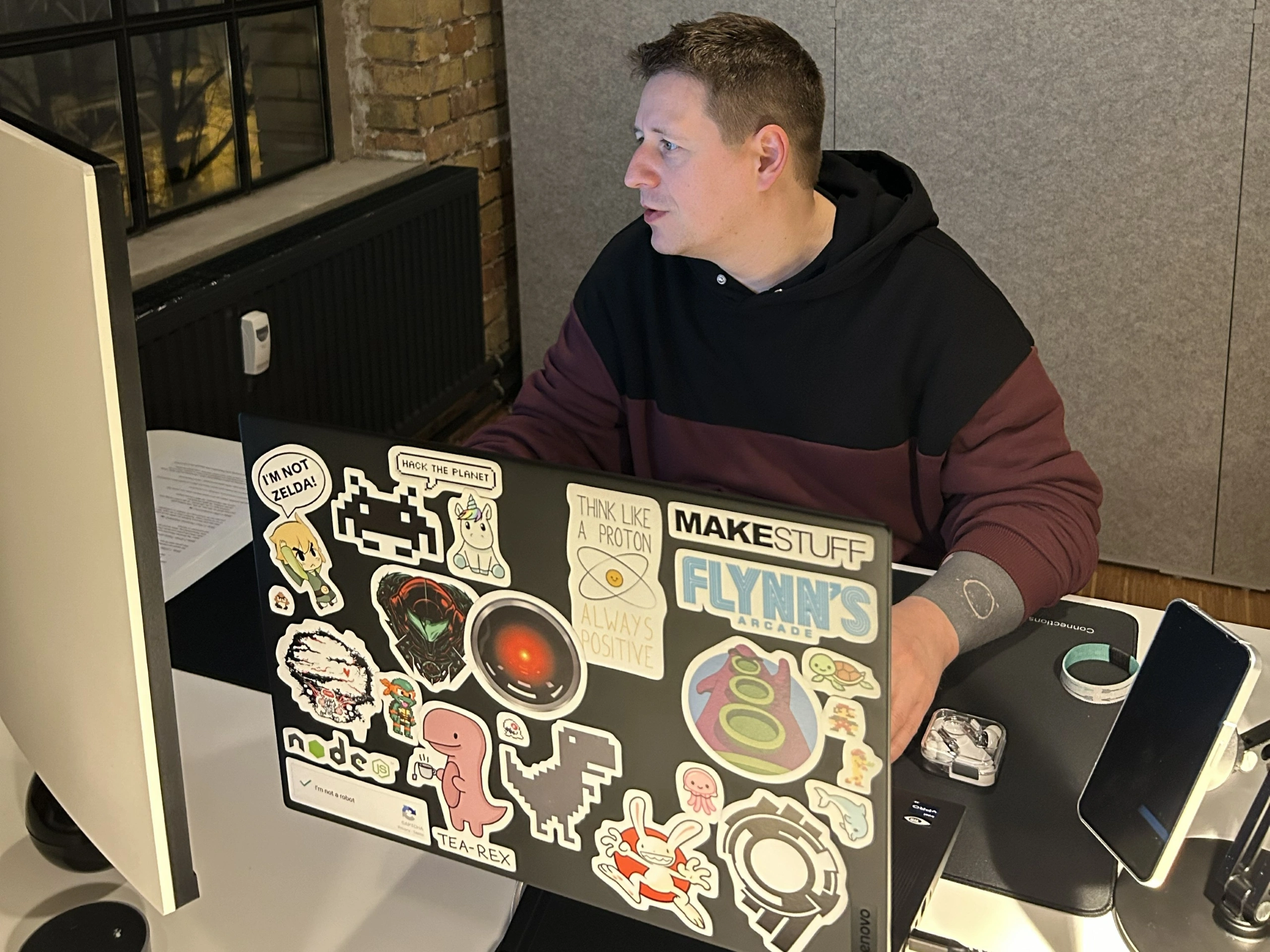 Ein Mann sitzt an einem Schreibtisch und arbeitet konzentriert an seinem Laptop. Die Rückseite des Laptops ist fast vollständig mit einer Vielzahl bunter Aufkleber bedeckt, darunter Motive wie ein T-Rex mit Teetasse ("Tea-Rex"), Sam & Max, Link ("I’m not Zelda"), das Node.js-Logo und ein "Flynn’s Arcade"-Sticker. Er trägt einen zweifarbigen Hoodie in Schwarz und Dunkelrot. Im Hintergrund sind eine rustikale Backsteinwand und ein Fenster zu sehen.