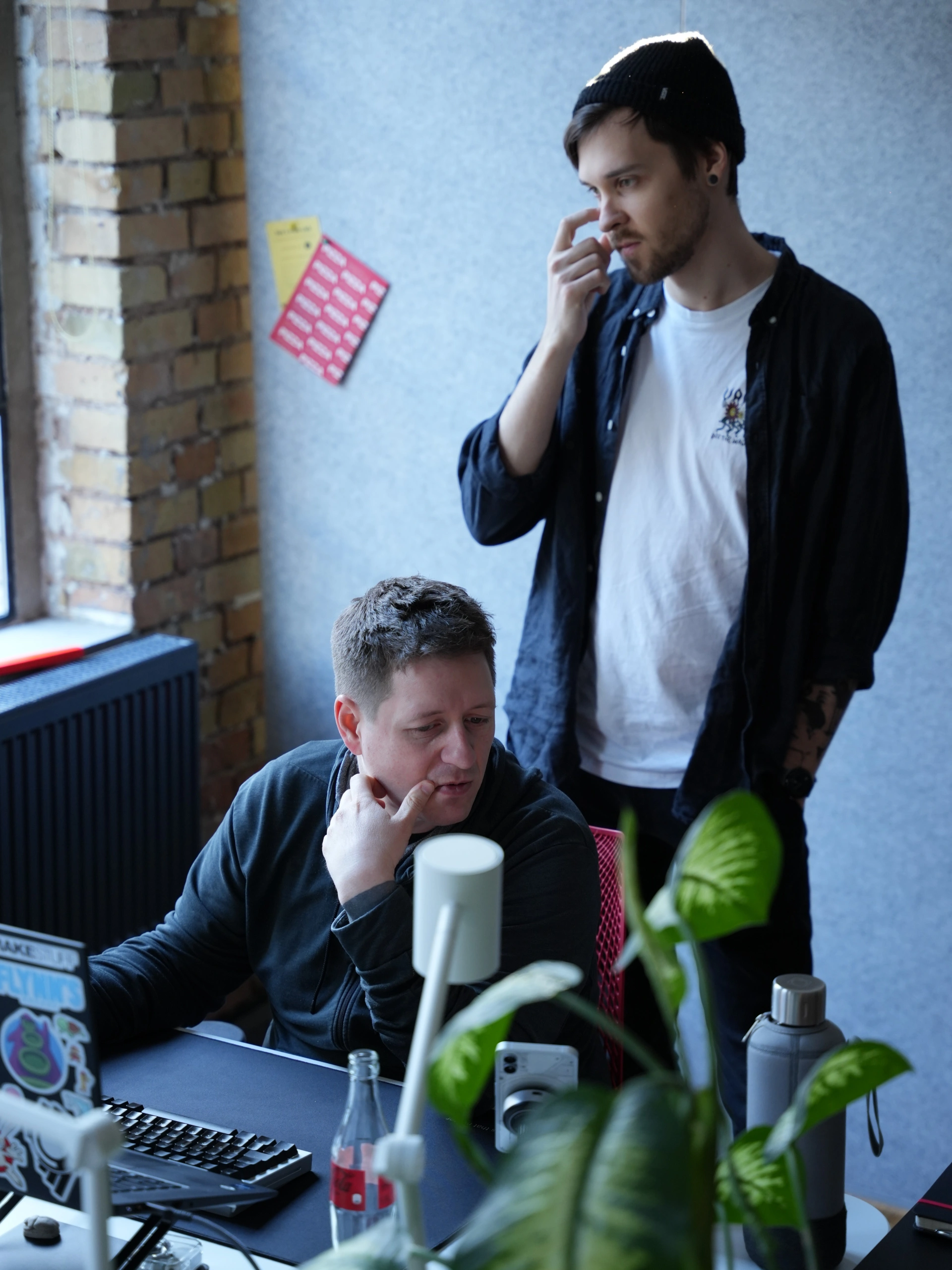 Zwei Männer in einer konzentrierten Brainstorming-Situation im Büro. Einer sitzt im Vordergrund am Schreibtisch, stützt nachdenklich sein Kinn auf die Hand und blickt auf einen Laptop. Der zweite Mann steht hinter ihm, trägt eine schwarze Beanie und blickt ebenfalls fokussiert nach unten. Im Hintergrund ist eine graue Akustikwand mit angepinnten roten Kärtchen zu sehen, im Vordergrund eine große Zimmerpflanze und eine Glasflasche auf dem Schreibtisch.