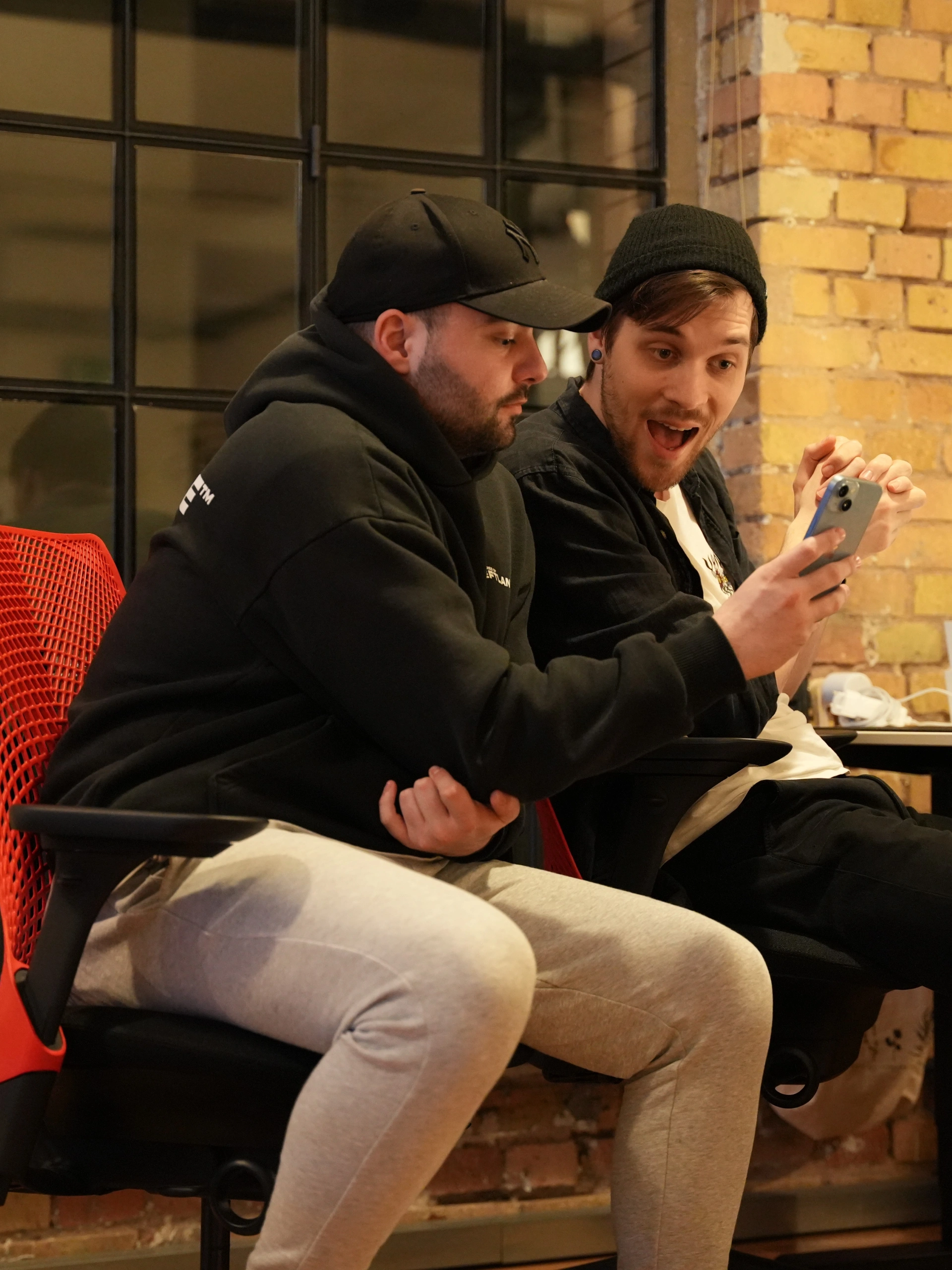 Zwei junge Männer sitzen nebeneinander in einem modern eingerichteten Raum mit Backsteinwand und großen Sprossenfenstern. Der Mann rechts trägt eine schwarze Beanie und blickt mit einem überraschten, lachenden Gesichtsausdruck auf ein Smartphone, das er in der Hand hält. Der Mann links, der eine schwarze Cap und einen dunklen Hoodie trägt, schaut ebenfalls konzentriert auf das Display. Beide sitzen auf ergonomischen Bürostühlen, wobei einer ein auffälliges rotes Netzdesign an der Rückenlehne hat.